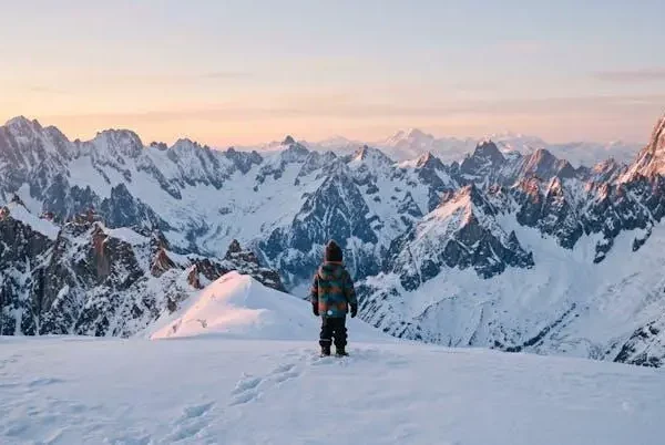 Bambino visto di spalle avvolto in un piumino colorato che osserva una catena montuosa innevata al sorgere del sole