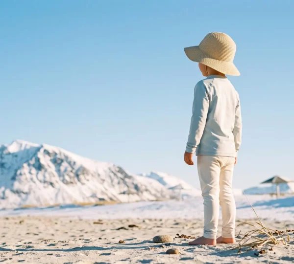 Bambino con cappellino e crema solare che gioca in un paesaggio che unisce simbolicamente montagna innevata e mare, protetto dal sole